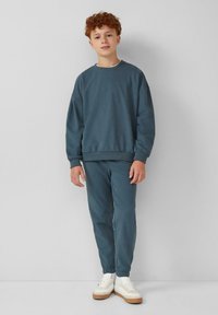 Ciemnoniebieska bluza i dopasowane dresy, z prążkowanymi mankietami i pasem. Model nosi białe sneakersy z brązowymi akcentami.