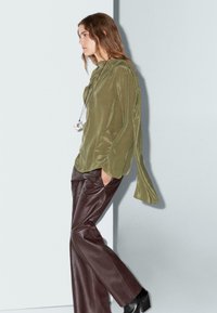 Blouse en soie vert olive avec des manches drapées, associée à un pantalon en cuir marron taille haute. Le mannequin porte un collier pendentif métallique.