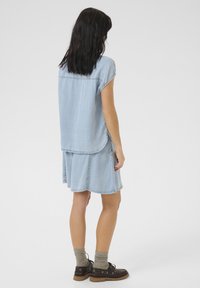 Abito di jeans azzurro chiaro con maniche corte, orlo arrotondato e design a strati. Abbinato a scarpe marroni e calzini grigi. Texture liscia.
