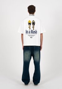 Un homme se tient de dos, portant un T-shirt blanc oversize avec un graphisme de deux cyclistes et le texte "In a Rush International", ainsi qu'un jean bleu délavé.