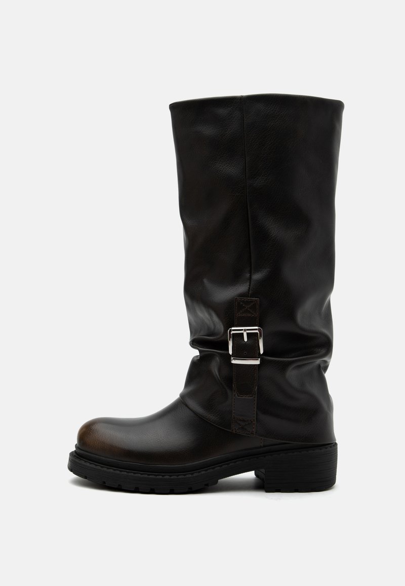 Botte haute en cuir noir avec bout arrondi, talon bas, bride à boucle latérale et tige légèrement relâchée sur fond blanc.