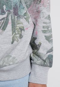 Sudadera gris con estampado de hojas tropicales coloridas con acentos en rosa, puños acanalados y textura suave.