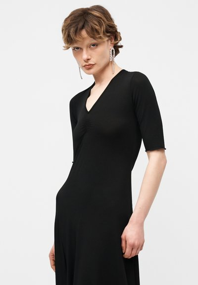 Άτομο με κοντά κυματιστά μαλλιά φοράει μαύρο εφαρμοστό φόρεμα με V-λαιμόκοψη και μανίκια μέχρι τον αγκώνα, καθώς και μακριά ασημένια σκουλαρίκια που κρέμονται.