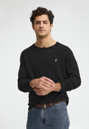 Homme aux cheveux bouclés portant un chemise noire à manches longues avec un petit logo blanc et un jean bleu, debout devant un fond clair uni.
