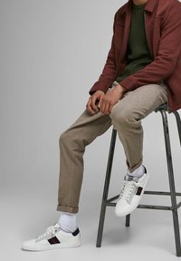 Białe trampki z granatowymi i czerwonymi akcentami, brązowe spodnie w teksturowaną kratę oraz zielony dzianinowy sweter pod bordową kurtką, siedzący na metalowym stołku.