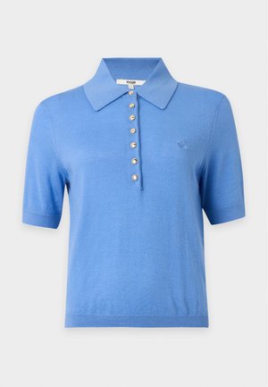 Kvinde iført en blå kortærmet polo og beige vidde bukser, der holder en brun læder skuldertaske med frynser.