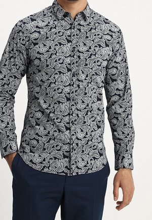 Mężczyzna w czarnej, zapinanej na guziki, długiej koszuli z misternym białym wzorem paisley oraz ciemnoniebieskich spodniach, ręce częściowo widoczne.