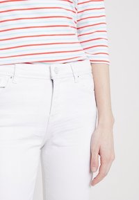 Jeans in denim bianco con design aderente e cuciture ben visibili. Indossati con una maglietta a righe con colori rosso, blu e bianco.