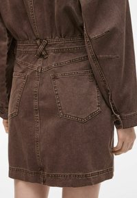 Smeđa traper haljina s povlačnom strukom, dva stražnja džepa i sašivenim detaljima. Tkanina ima blago teksturirani izgled.