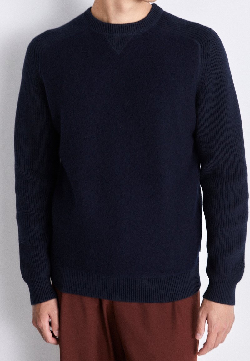 Homme portant un pull en maille bleu marine avec poignets et ourlet côtelés, associé à un pantalon marron, sur un fond clair uni.