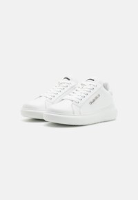 White leather sneakers with flat laces, rounded toe, and a textured sole. Features a silver logo on the side and padded collar.