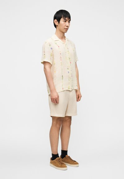 Μπλούζα από λινό σε μπεζ χρώμα με κοντά μανίκια και κεντήματα με λουλουδάτα σχέδια, σε συνδυασμό με σορτς σε ανοιχτό μπεζ χρώμα. Ολοκληρώνουν το σύνολο καφέ σουέντ αθλητικά παπούτσια και μαύρες κάλτσες.