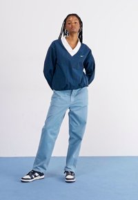 Navy pullover with white collar, loose fit, paired with light blue jeans and black sneakers with white accents, standing on a blue surface.