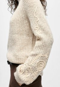 Bēšs adīts džemperis ar teksturētu virsmu, rotāts ar smalkām krēmkrāsas izšuvēm gar piedurknēm un ribotām apmalēm.