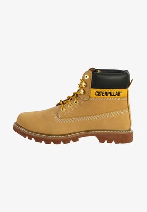 Bottine en cuir marron avec un col noir, des accents jaunes, des œillets dorés et une semelle robuste. Présente une couture latérale et un détail de surpiqûre contrastante.