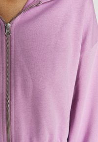 Veste mauve clair zippée en tissu doux et extensible, avec une fermeture éclair en métal et une coupe décontractée, idéale pour un usage quotidien.