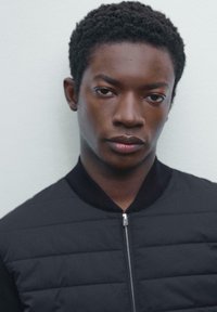 Giubbotto bomber nero con zip e design imbottito. Presenta un colletto a coste e una texture sottile. Il modello mostra un'espressione neutra.