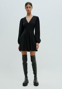 Schwarzes Mini-Kleid mit langen Puffärmeln, V-Ausschnitt und vorderen Knöpfen. Trägt hohe schwarze glänzende Stiefel bis zum Knie. Glatte Stofftextur.