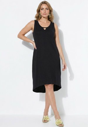 Femme portant une robe noire sans manches avec une petite découpe au niveau du décolleté, des boucles d'oreilles vertes et des sandales jaunes à fleurs, debout contre un mur blanc.