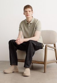 Beige polo skjorte med korte ermer, mørke jeans og lyse ankelstøvler. Sitter på en lys, polstret stol med trebein.