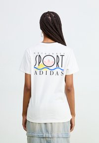 Valkoinen puuvillainen T-paita, jossa pyöreä kaula-aukko ja monivärinen graafinen kuvio selässä, sisältäen sanan "SPORT" ja Adidas-logon.