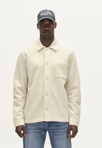 Krémová vlněná bunda s knoflíkovým zapínáním a jednou náprsní kapsou. V kombinaci s modrými džínami a tmavě zelenou kšiltovkou s vyšitým textem.