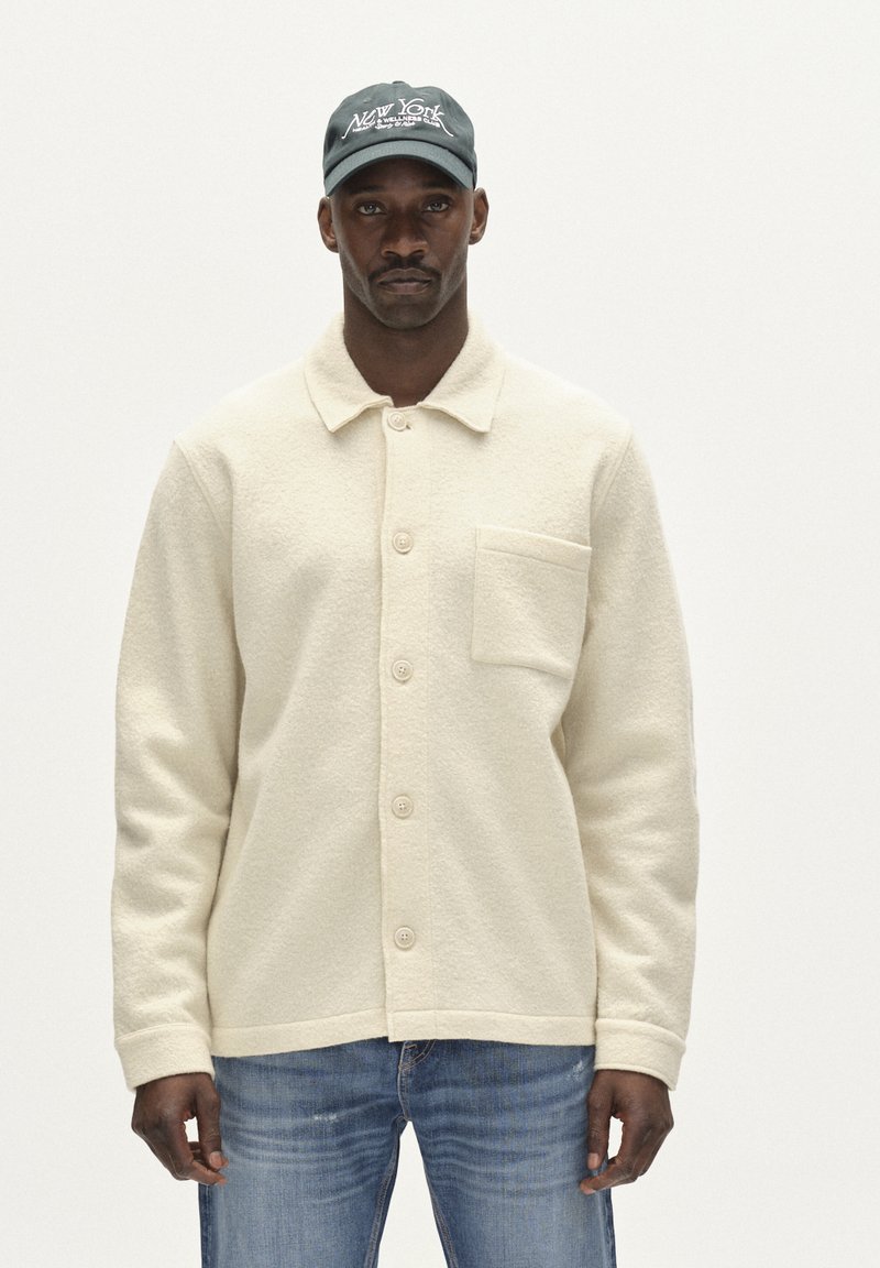 Krémová vlněná bunda s knoflíkovým zapínáním a jednou náprsní kapsou. V kombinaci s modrými džínami a tmavě zelenou kšiltovkou s vyšitým textem.