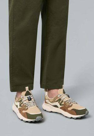 Beige und grüne Multimaterial-Sneaker mit einer strukturierten Oberseite, gepolsterter Schuhkragen und weißer Sohle. Merkmale sind auffällige Schnürung und Markenlogo.