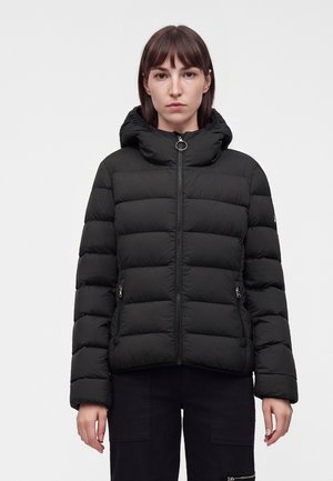 Frau mit braunen Haaren trägt eine schwarze Steppjacke mit hohem Kragen und Frontreißverschluss und steht vor einem schlichten hellen Hintergrund.
