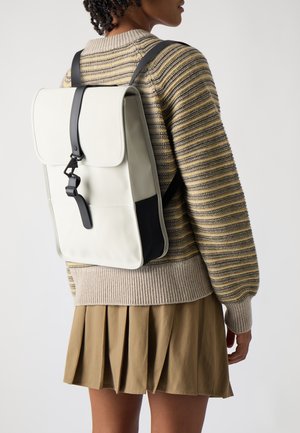 Person wearing a cream and black rectangular backpack with a front buckle over a striped sweater and pleated tan skirt.