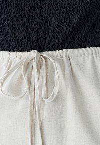 Melnas kroku auduma augšdaļa ar dabīgas krāsas, teksturētu auduma aukliņu jostu. Raksturīga priekšējā mezgla detaļa un redzamas šuves.