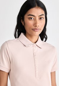 Camisa polo em algodão rosa claro, com gola clássica, fecho de botão e logótipo bordado discreto no lado esquerdo do peito.