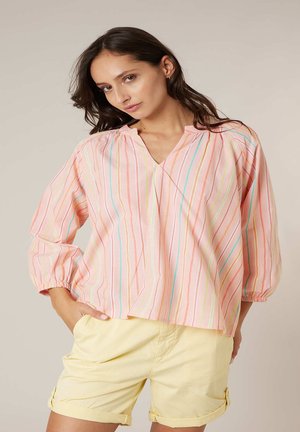 Vrouw draagt een loszittende roze gestreepte blouse met lange mouwen en lichtgele opgerolde korte broek, staand met één hand in de zak.