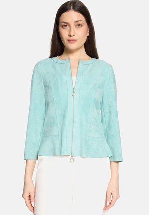 Kvinde iført lyseblå lynlåstrøje med 3/4-ærmer og hvide bukser, stående foran en ensfarvet hvid baggrund.