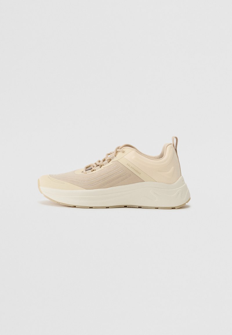 Chaussure de sport beige avec un dessus lisse et texturé, dotée d'une semelle rembourrée et de fines rayures réfléchissantes. Terminée avec une languette de tirage.