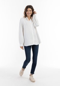 Witte blouse met lange mouwen, relaxte pasvorm en gebogen zoom, in combinatie met donkerblauwe slim fit jeans en beige sneakers.