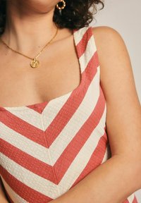 Abito a righe rosse e crema con scollatura quadrata, tessuto texturizzato e dettaglio a cucitura triangolare, abbinato a una collana dorata.