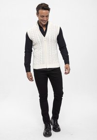 Elfenbeinfarbene Strickweste mit Kabelmuster, mit Reißverschluss vorne und ohne Ärmel. Kombiniert mit einem schwarzen Langarmshirt und schwarzen Hosen, abgerundet mit schwarzen Schuhen.