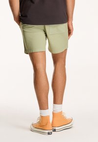 Svetle zelené šortky s kapsou na zadní straně, spárované s oranžovými vysokými teniskami a bílými ponožkami do kotníků. Jednoduchý, neformální design.