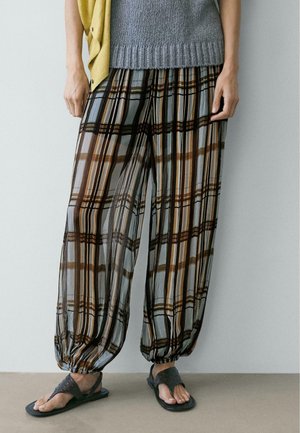 Personne portant un pantalon ample à carreaux avec des poignets froncés, un haut en tricot gris, un gilet jaune et des sandales noires à bout ouvert, se tenant contre un mur uni.