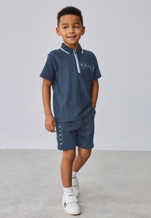 Ung dreng står, iført navyblå kortærmet polo og matchende shorts med hvide sneakers, smilende og kigger til siden.