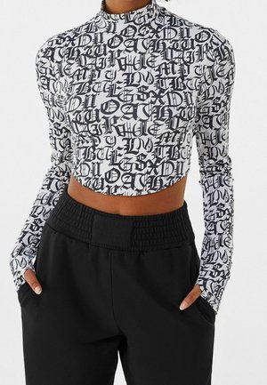 Femme portant un crop top blanc à manches longues avec des lettres en écriture gothique noire et un pantalon noir taille haute, les mains dans les poches.