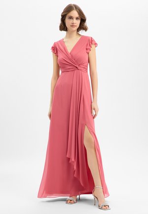 Vrouw die een vloerlange roze jurk draagt met geplooide mouwtjes, een wikkelcorset, een hoge split en zilveren sandalen met hoge hakken, staand tegen een witte achtergrond.