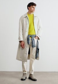 Homme portant un trench-coat beige, un pull jaune, un pantalon blanc, une chemise grise à carreaux nouée à la taille, et des bottes grises, debout devant un fond blanc uni.