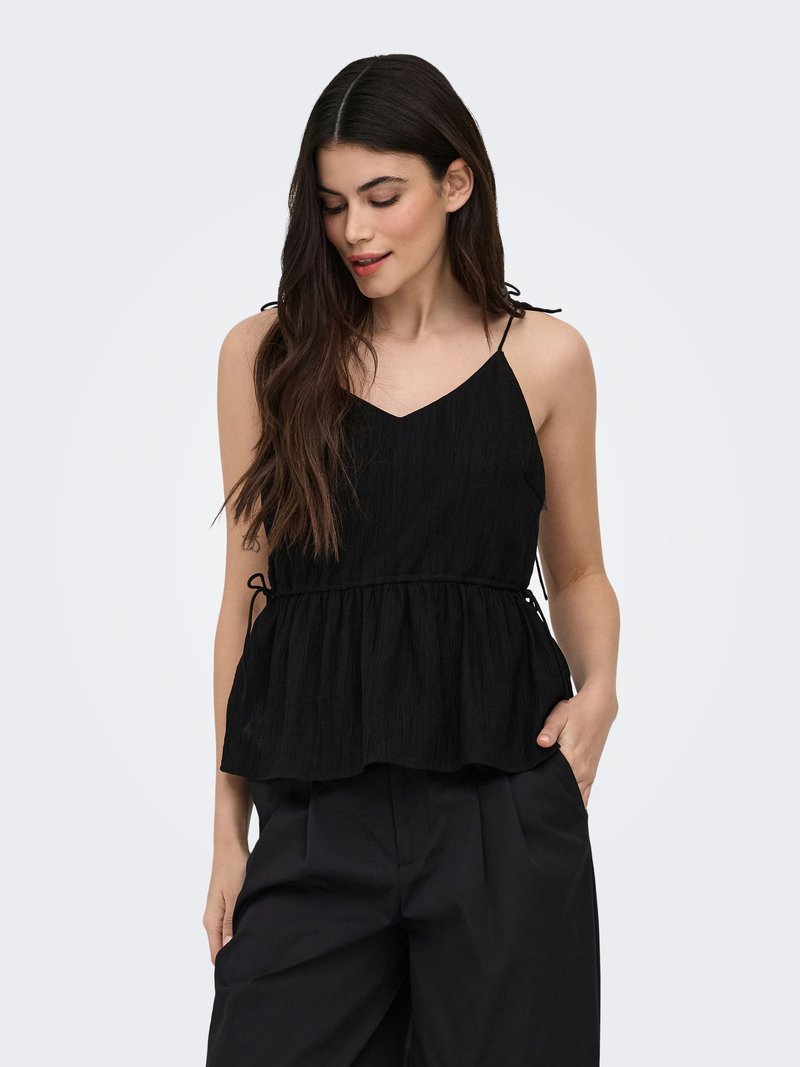 Femme aux longs cheveux foncés portant un top peplum noir sans manches et un pantalon noir à jambes larges, debout, une main dans la poche.