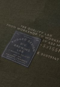 Kangasmarmori oliivinvihreä kangas, jossa on teksturoitu tummansininen etiketti, jossa on valkoista tekstiä "RUGGED DESIGN DIV. BREWING LAB" ja erilaisia tuotedetalleja.