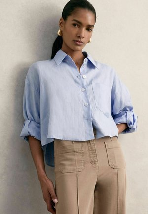 Világoskék, rövidített gombos ing gallérral, felgöngyölt ujjal és mellzsebbel, passeggerírozott bézs nadrággal, amely hátul zsebekkel rendelkezik.