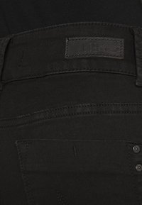 Černé džíny se hladkým povrchem, s koženou visačkou označenou "LTB" a kovovými hroty na zadní kapse pro zvýraznění detailu.