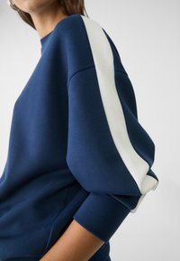 Felpa blu navy con dettagli a strisce bianche sulle maniche; presenta un collo rotondo e polsini aderenti, realizzata in morbido tessuto testurizzato.