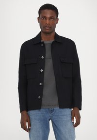Schwarze Leinenjacke mit zwei Brusttaschen, Knopfverschluss und Kragen. Tragen über einem grauen Hemd und kombiniert mit hellblauen Jeans.